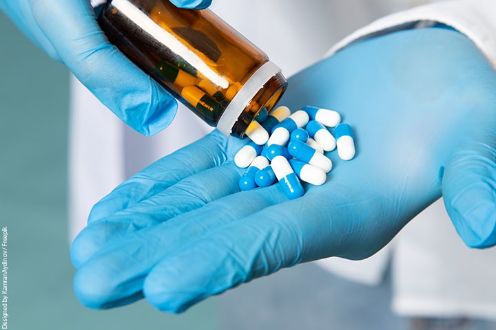 Um vidro de pílulas azuis e brancas, as quais caem na mão de uma pessoa com luvas