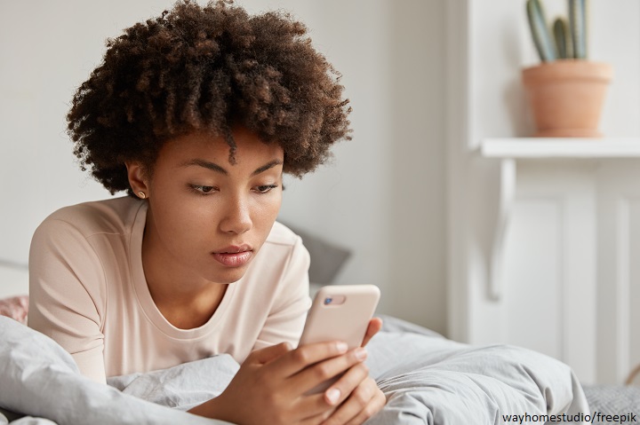 image of black young lady with afro haircut checks new update on social networks shares multimedia files on mobile phone dressed in casual nightclothes rests in bed makes payment online