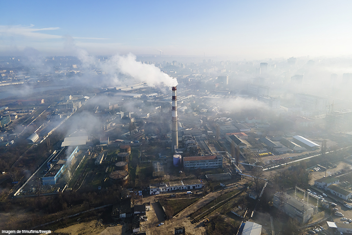 Cidade vista do alto com vários pontos de fumaça na atmosfera; ao centro da foto, chaminé de uma fábrica se destaca